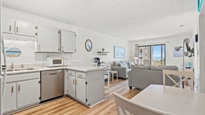 A modern kitchen with white cabinets, a microwave, and a dishwasher. There is a dining area with a table and chairs, and a gray sofa in the living room.