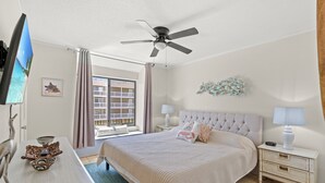 A hotel room with a bed, a TV, a ceiling fan, a window with curtains, and a view of a building outside.
