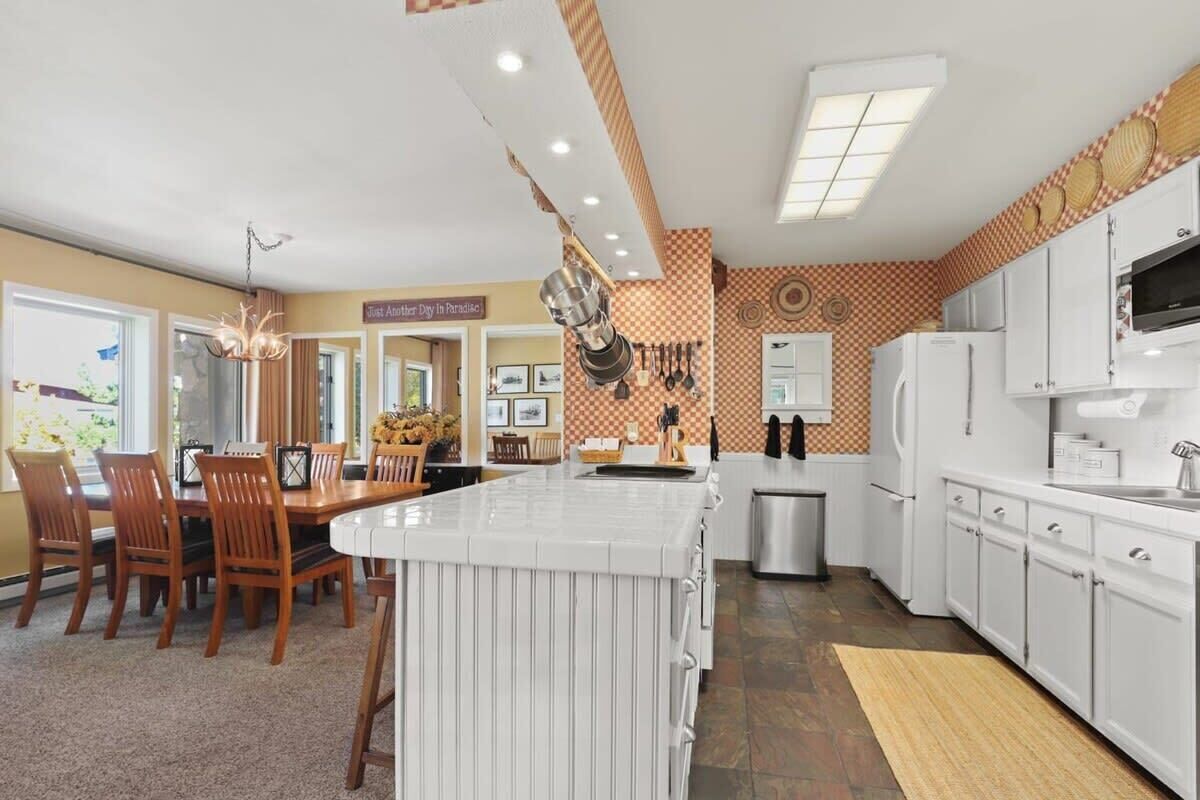 Une cuisine moderne avec des meubles blancs, un îlot central et un coin repas agrémenté de chaises en bois et d’un lustre.
