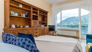 Une chambre d’hôtel dotée d’une grande armoire en bois, d’un lit avec des coussins à motifs, d’une télévision et d’un balcon offrant une vue sur la montagne et la mer.