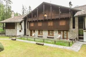A two-story wooden house with stone walls, a balcony, and a stone pathway leading to the entrance.