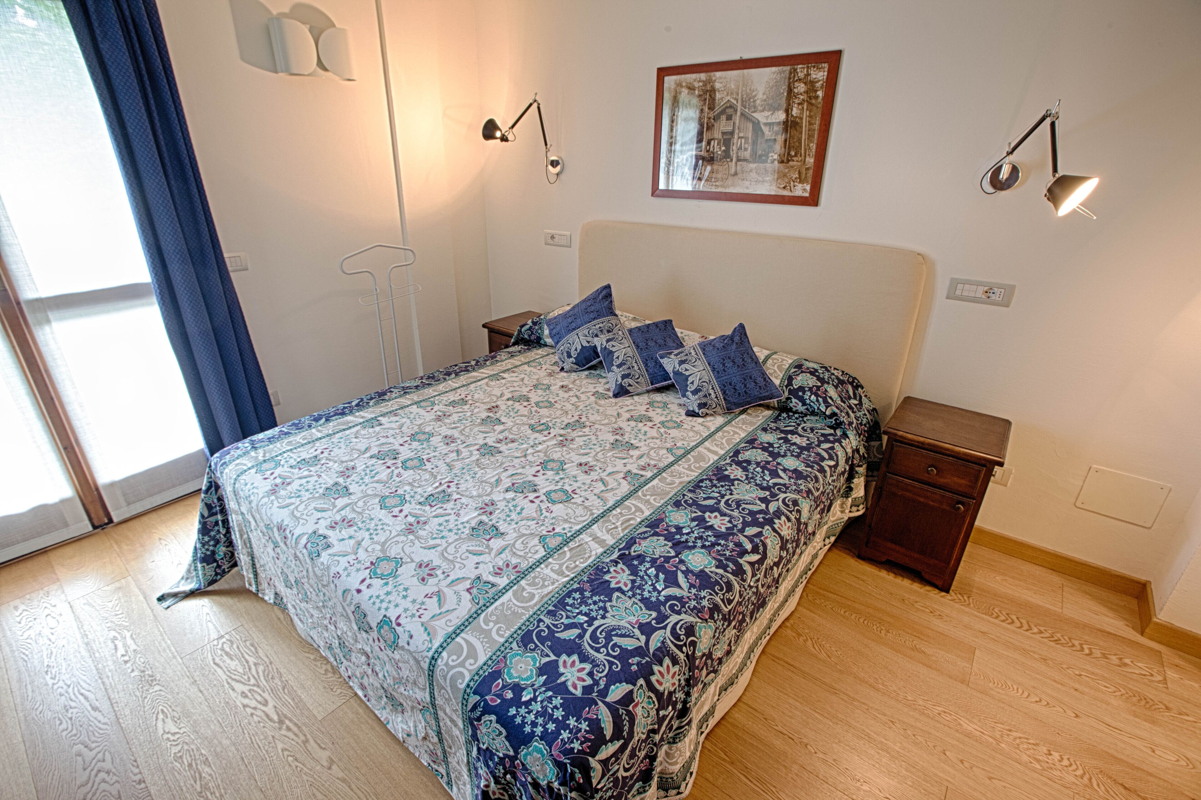Un dormitorio con una cama grande, dos mesitas de noche, una lámpara de pared y un ventanal con cortinas.
