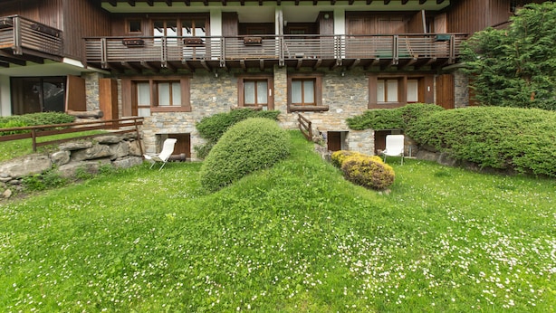 A multi-story wooden building with balconies, stone walls, and a well-maintained garden with white flowers. - JOIVY Lovely Apt W/Mountain View And Parking In Courmayeur (Courmayeur)