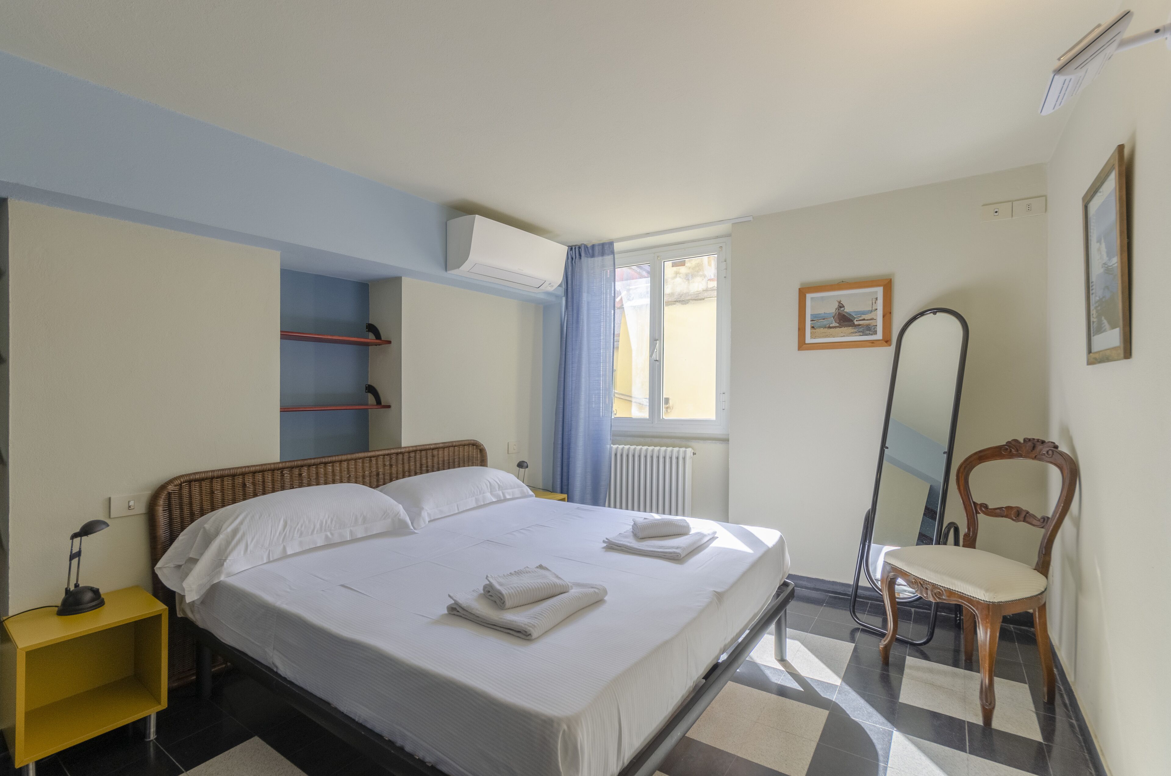 Un lit bien fait, avec du linge de lit et des serviettes blanches, une chaise en bois, un miroir et une étagère dans une pièce dont le sol est recouvert de carreaux.