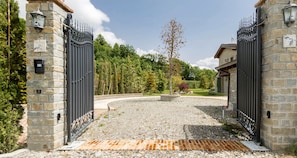 A gated entrance with stone walls and metal gates, leading to a gravel driveway and a landscaped area with trees and shrubs. - JOIVY Superb Villa For 7 W/Backyard And Bbq Area In Vaccarezza (Bobbio)
