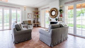 A spacious living room with two grey sofas, a fireplace, and large windows offering a view of greenery outside. - JOIVY Superb Villa For 7 W/Backyard And Bbq Area In Vaccarezza (Bobbio)