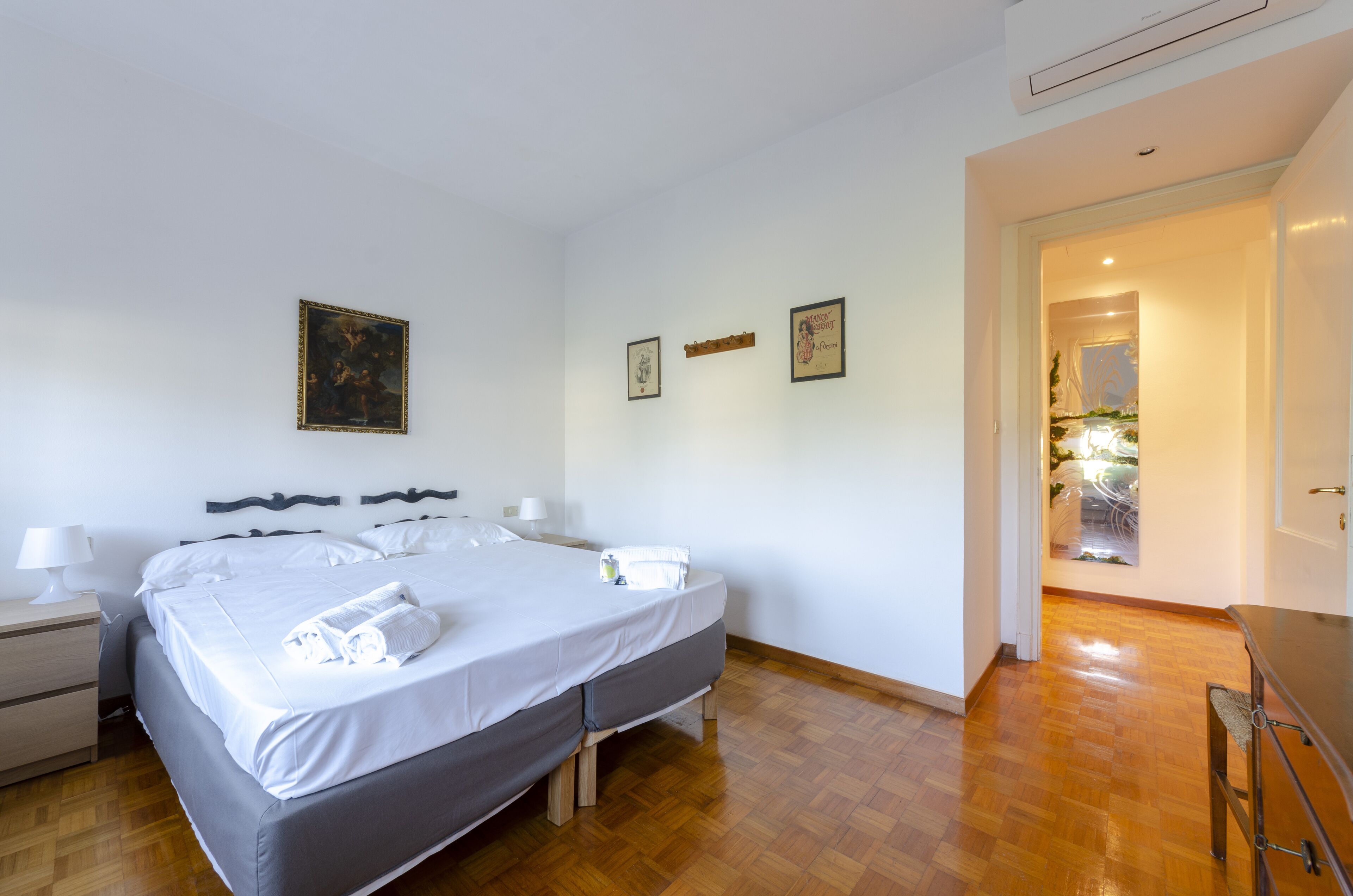 A neatly made bed with white linens and a gray headboard, a wooden nightstand with a lamp, and a wooden floor.