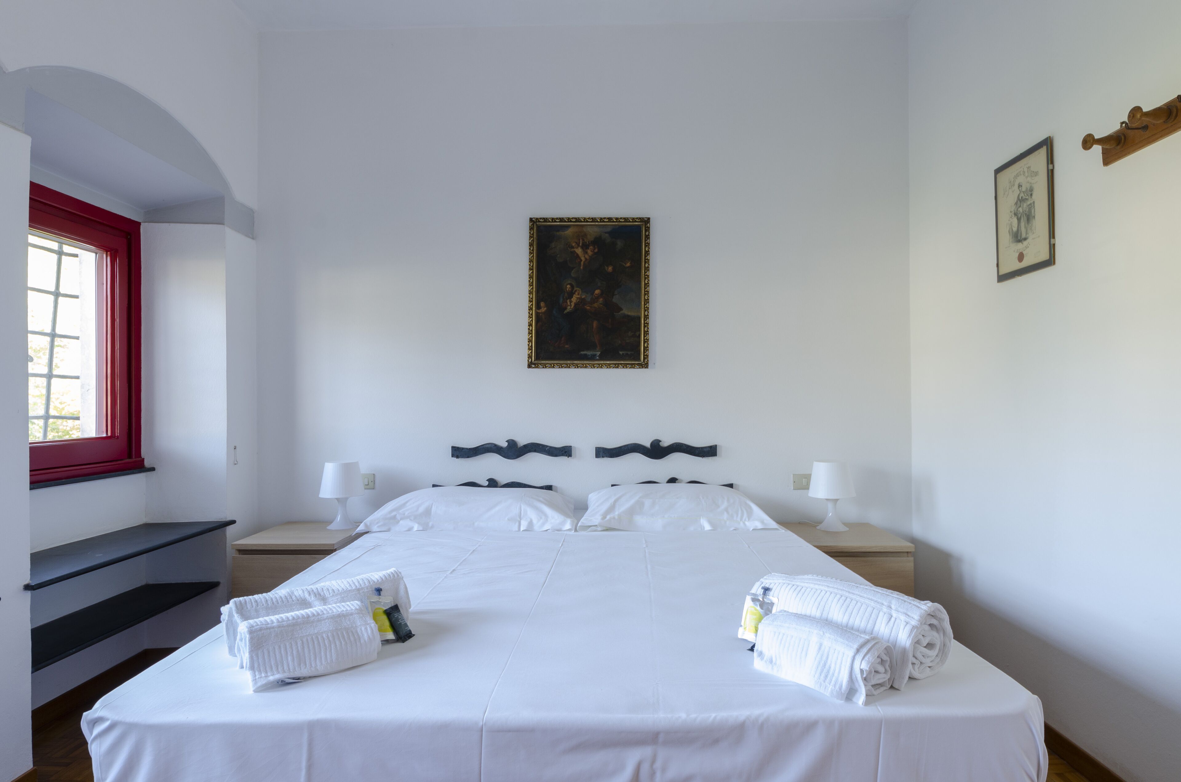 A hotel room with a large bed, two bedside tables with lamps, a painting on the wall, and a window with a red frame.