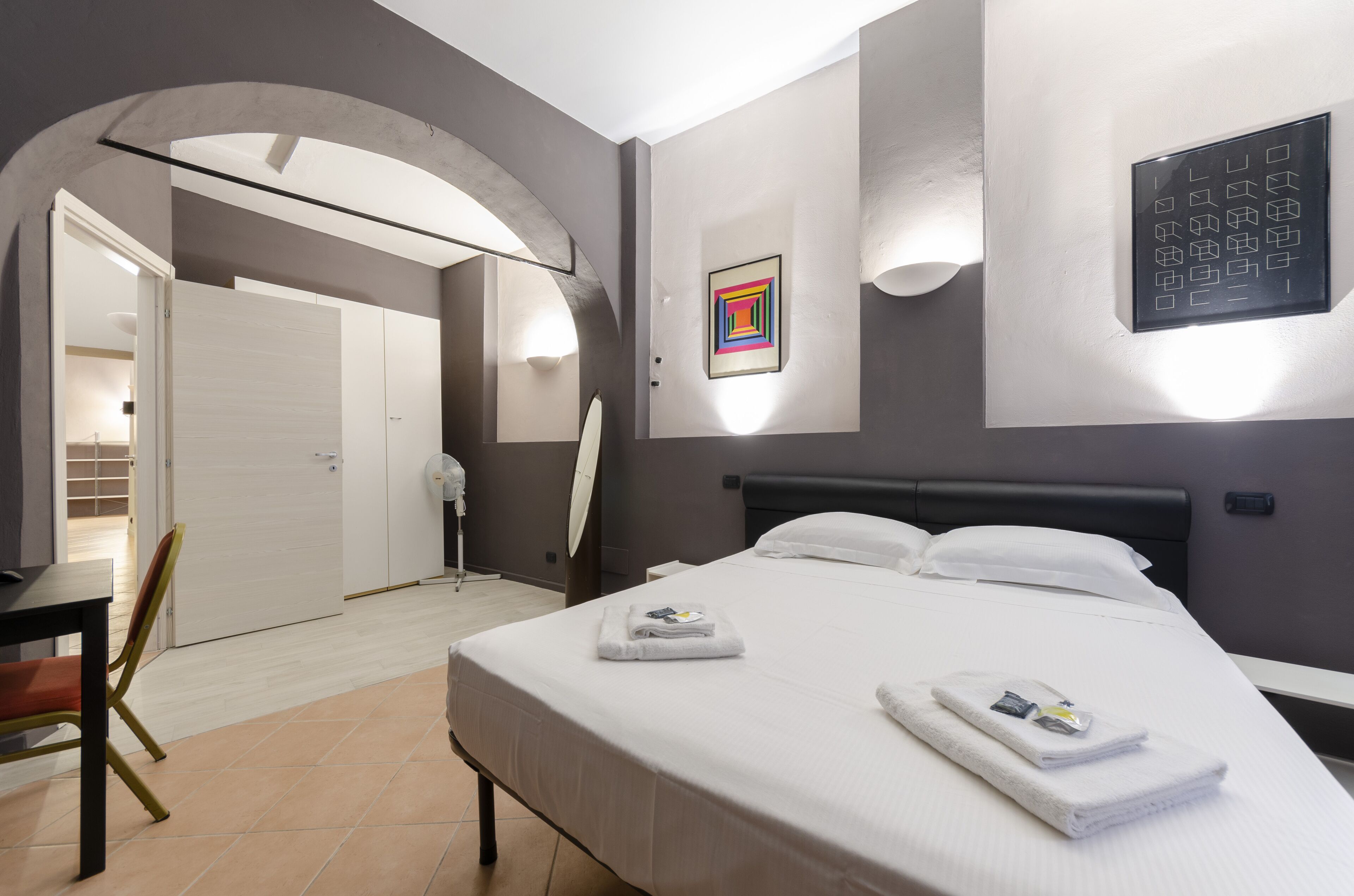 A modern hotel room with a bed, a desk, and a chair. There is a door leading to another room and a wall-mounted light fixture.