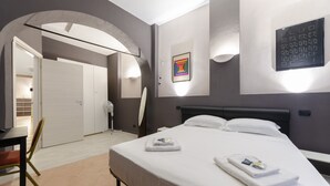 A modern hotel room with a bed, a desk, and a chair. There is a door leading to another room and a wall-mounted light fixture. - JOIVY Warm Family Flat For 9, Near Porto Antico (Genoa)