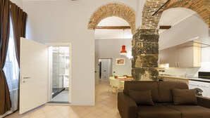 A modern living room with a brown sofa, a dining area with a table and chairs, and a kitchen with a stone column. - JOIVY Warm Family Flat For 9, Near Porto Antico (Genoa)