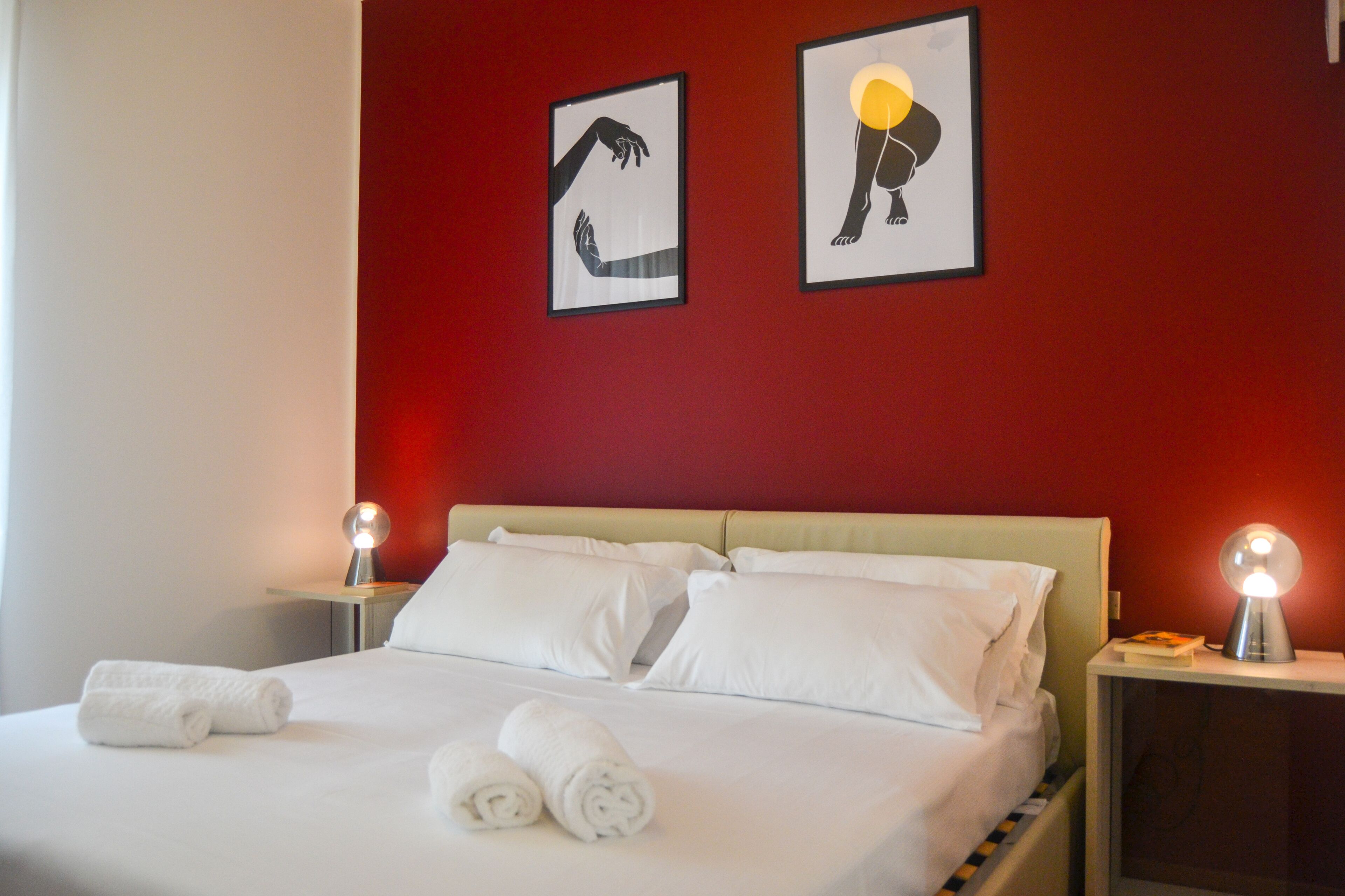 A neatly made bed with white linens and two rolled towels, a red wall with two framed artworks, and bedside lamps on nightstands.