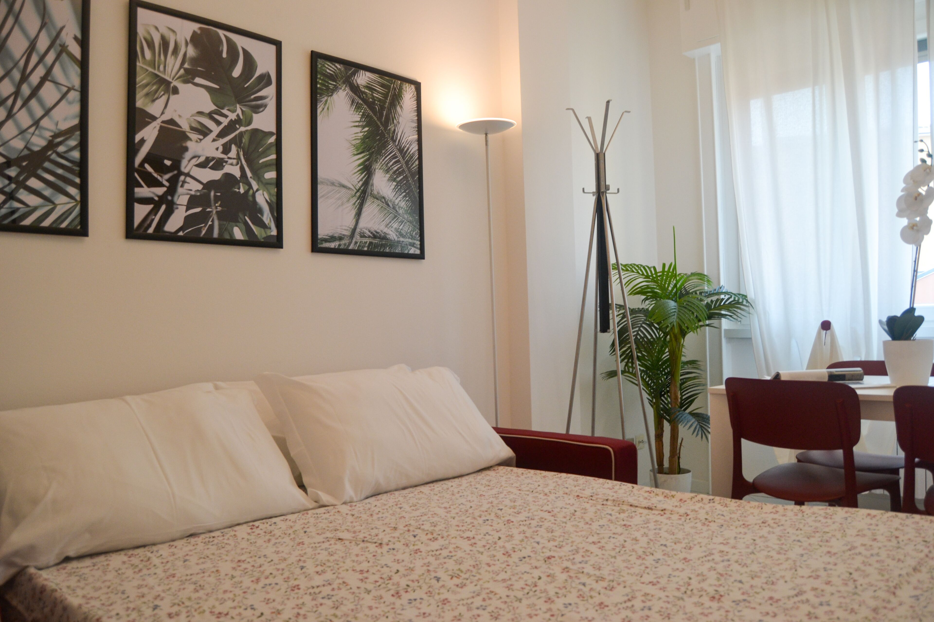 A bedroom with a bed, a desk, and chairs. There are two framed pictures on the wall, a floor lamp, and a potted plant.