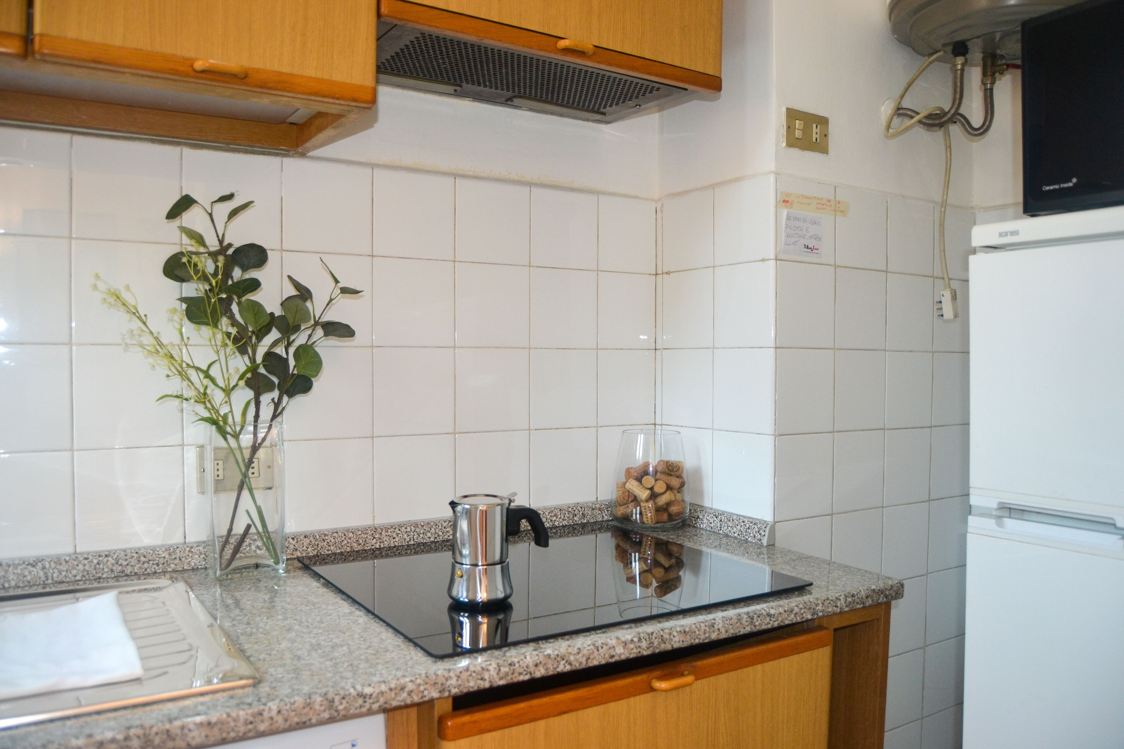 A kitchen with white tiles, a stainless steel coffee pot, a glass jar with corks, and wooden cabinets.