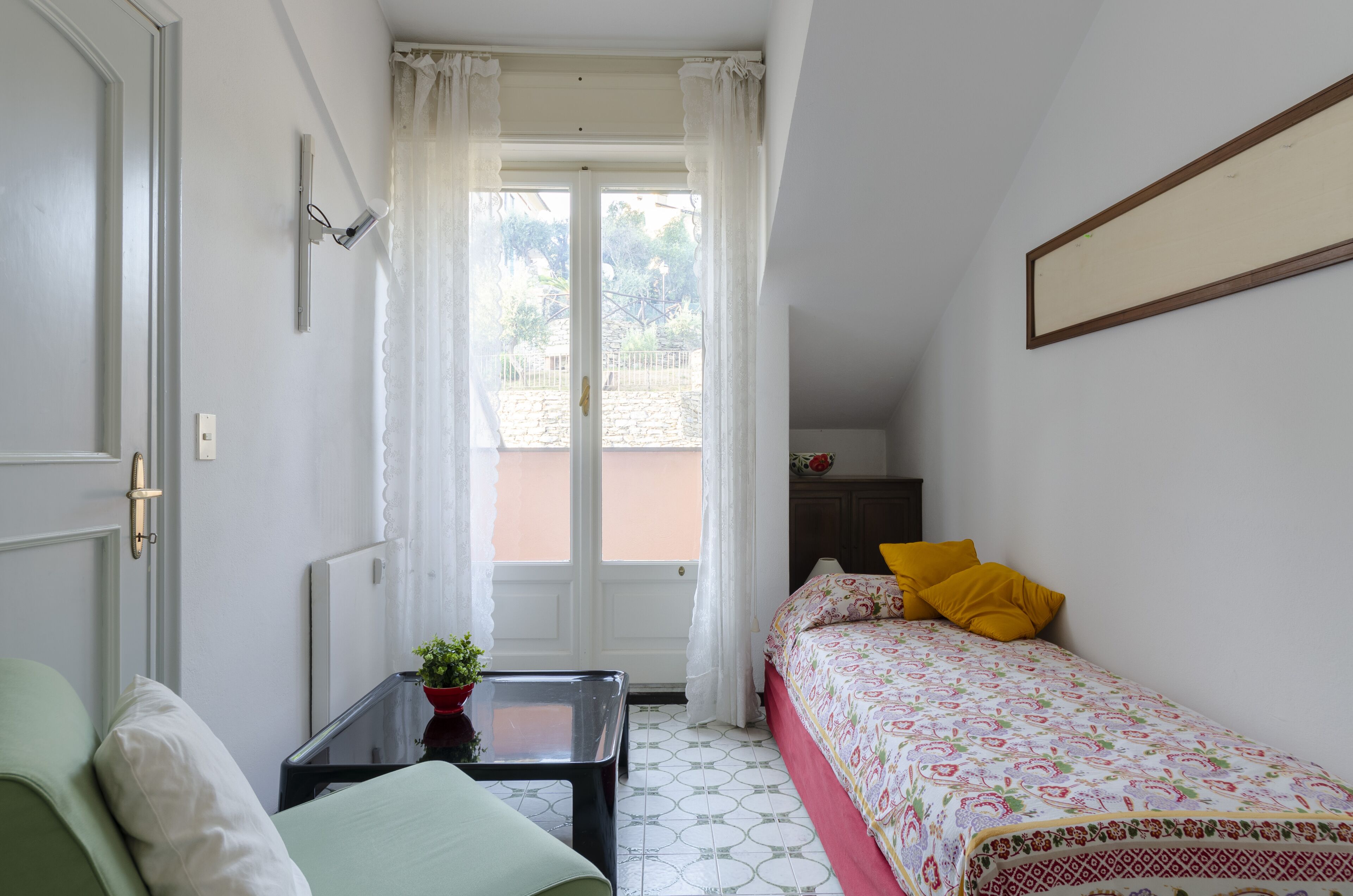 Ein kleiner, gut beleuchteter Raum mit einem Bett, einem Tisch und einem Fenster mit Vorhängen.