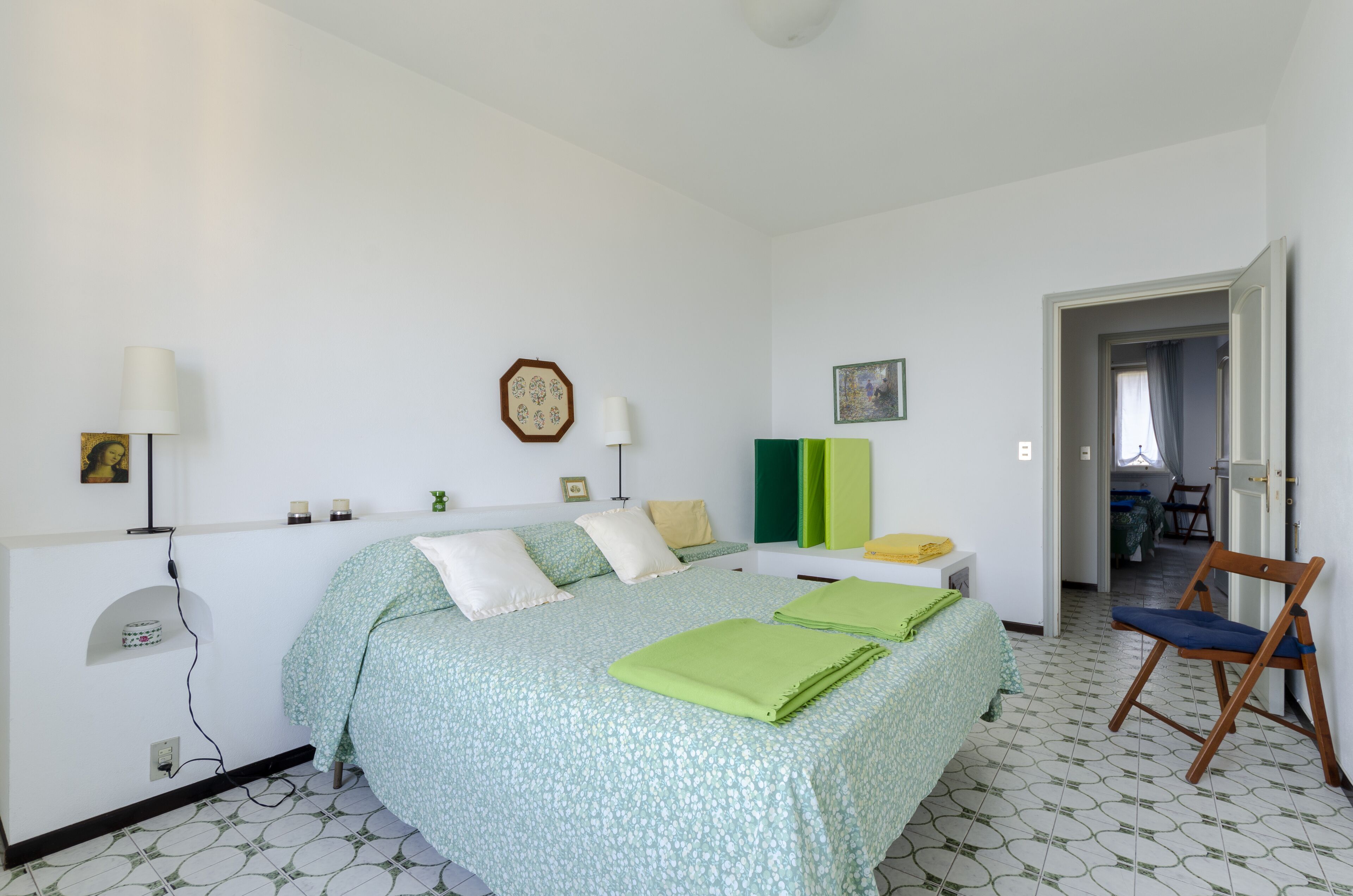Ein Schlafzimmer mit Bett, Nachttischlampen, einem Sessel und einer Tür zu einem anderen Raum.