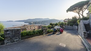 Une route côtière bordée de murs de pierre, où sont garés des scooters, et qui offre une vue sur la mer et les collines lointaines.