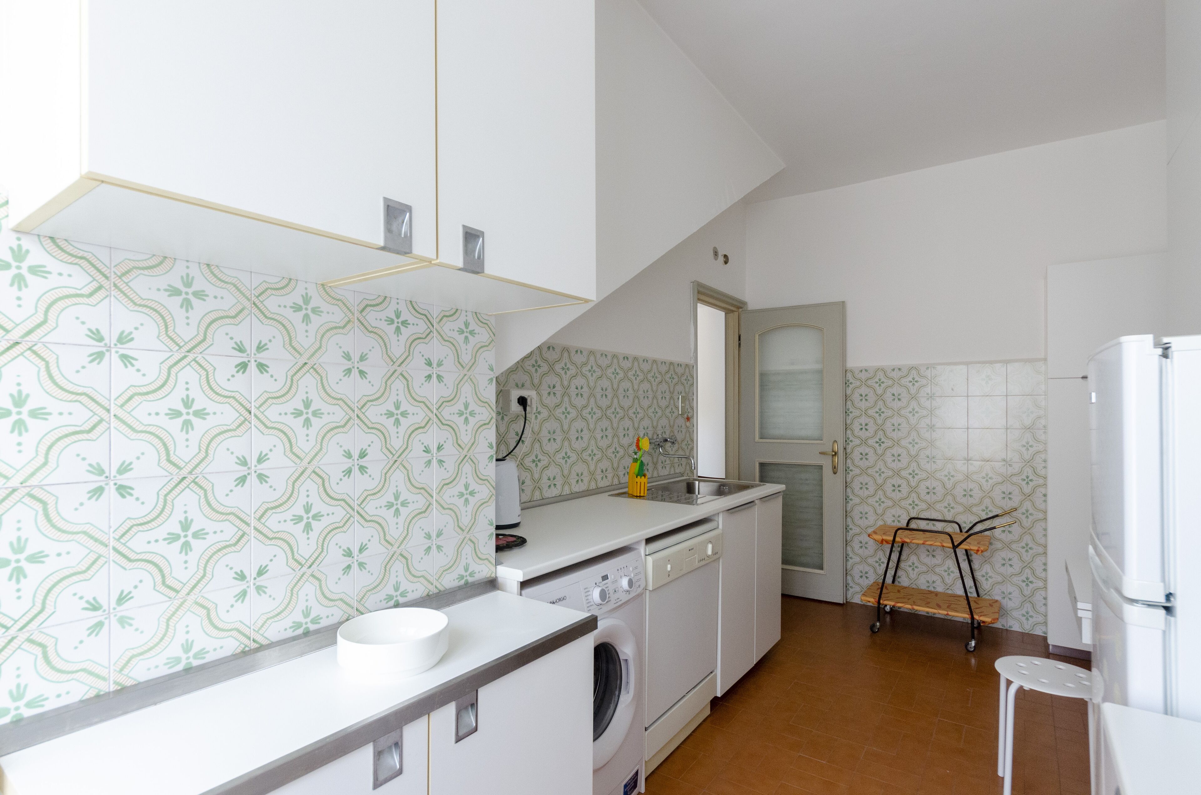 A compact kitchen with white cabinets, a washing machine, a sink, and a small cart.