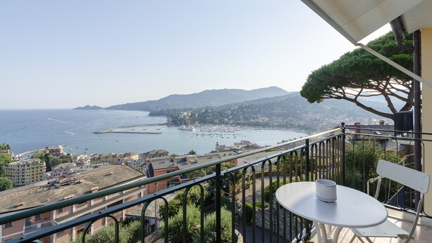Un balcon avec une table et une chaise, donnant sur une ville côtière avec ses bâtiments et offrant une vue sur la mer et les montagnes.