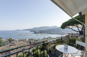 A balcony with a table and chair, overlooking a coastal town with buildings and a view of the sea and mountains. - JOIVY Spectacular Sea View Apt For 5 W/Terrace (Rapallo)