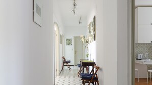 Un couloir étroit mène à un coin repas comprenant une table et des chaises, une porte blanche et un espace cuisine avec une bouilloire et un meuble blanc.