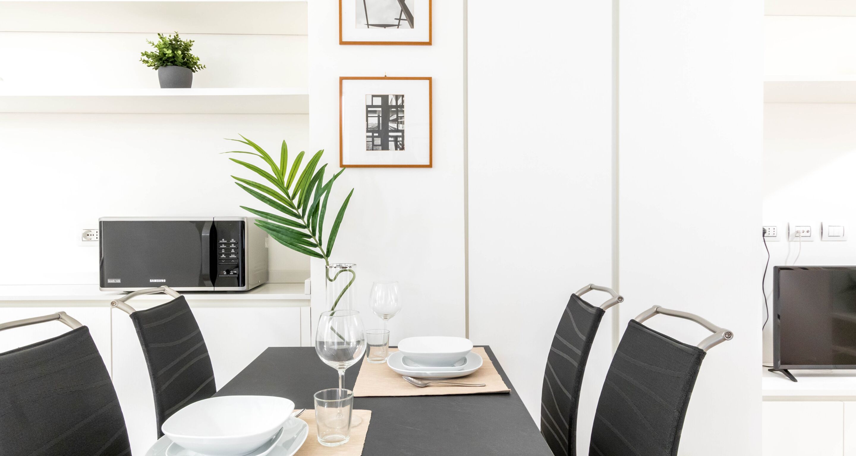 A modern dining area with a black table, white plates, and black chairs. There is a potted plant, a framed picture, and a microwave on the wall.