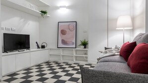 Een moderne woonkamer met een zwart-wit geruite vloer, een grijze bank met rode kussens, een flatscreen televisie, een lijst met een abstract schilderij en een vloerlamp.