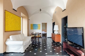 A hotel room with a white sofa, a dining table with chairs, a flat-screen TV, and artwork on the walls.