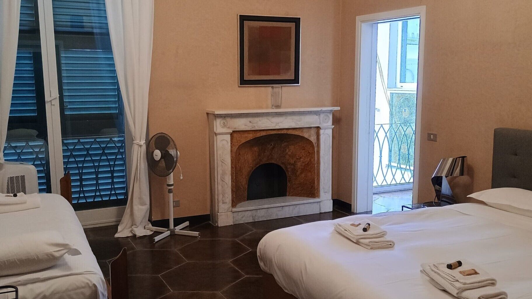 A hotel room with two beds, a fireplace, a standing fan, and a view of the outside balcony.