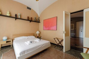 A hotel room with a bed, bedside table, lamp, and a shelf with decorative items.