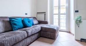 A grey sectional sofa with two blue cushions, a radiator, a sliding glass door, and a potted plant. - JOIVY Elegant 1 Br Flat Next To Milano Centrale (Milan)