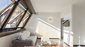 A modern room with a sofa, a dining table with chairs, a TV mounted on the wall, and large windows allowing natural light.