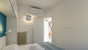 A hotel room with a bed, a radiator, a wall-mounted air conditioner, and a door leading to another room.