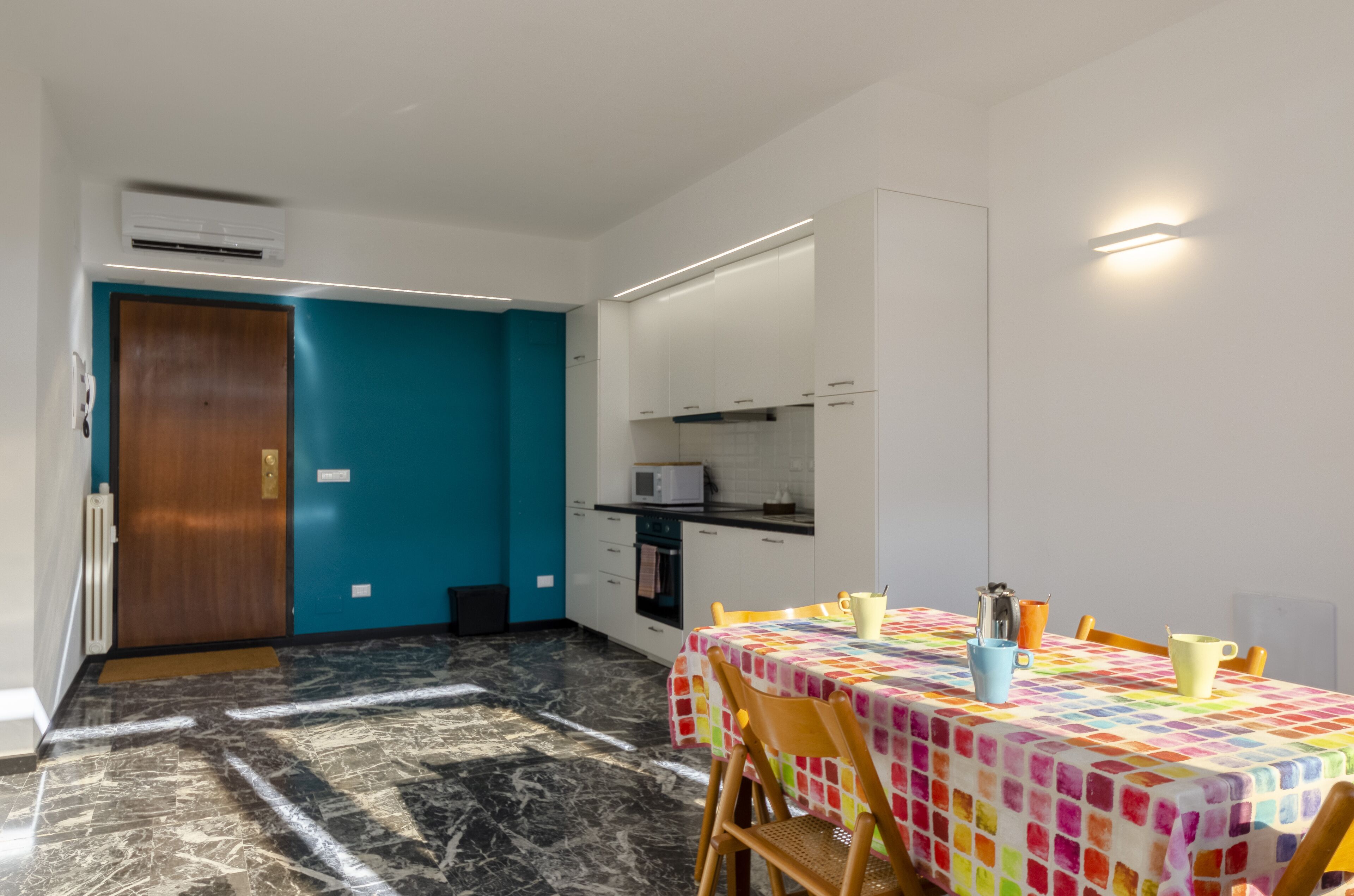Una cocina compacta con armarios blancos, un microondas y un lavabo. Hay un comedor con una mesa cubierta por un mantel colorido y sillas.