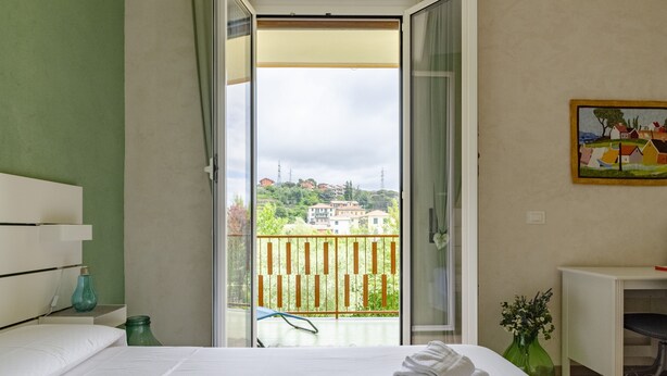 A hotel room with a bed, a small table, a chair, a painting on the wall, and a balcony with a view of buildings and greenery. - JOIVY Chic Chiavari Apt With 2 Spacious Terraces (Chiavari)