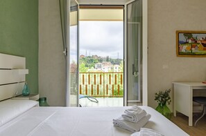 A hotel room with a bed, a small table, a chair, a painting on the wall, and a balcony with a view of buildings and greenery. - JOIVY Chic Chiavari Apt With 2 Spacious Terraces (Chiavari)