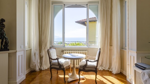 A room with a round table and two chairs, a radiator, and a view of the sea through a large window with curtains. - JOIVY Splendid Villa With Orange Trees And Stunning View (Santa Margherita Ligure)