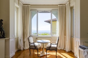 Une pièce avec une table ronde et deux chaises, un radiateur et une vue sur la mer par une grande fenêtre avec des rideaux.