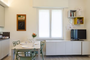 Une cuisine compacte comprenant une table à manger, des chaises, un petit îlot central agrémenté d’un vase de fleurs, un téléviseur posé sur un meuble et une bibliothèque contenant des livres et des objets décoratifs.