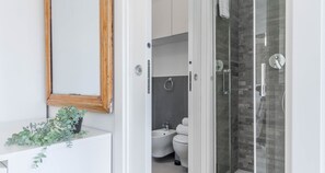 Et moderne badeværelse med en hvid vask, et spejl med træramme, en potteplante, en brusekabine i glas og et bidet.