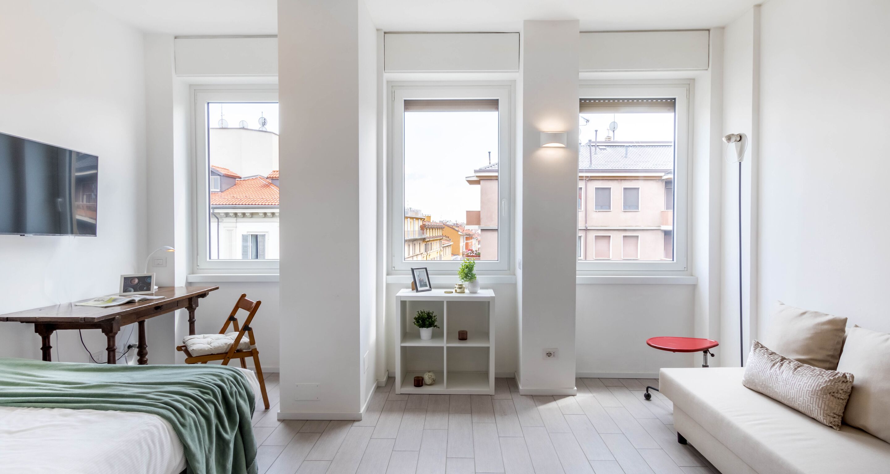 A modern, well-lit room with a bed, a desk, a small table, and a sofa. There are large windows providing a view of the cityscape.