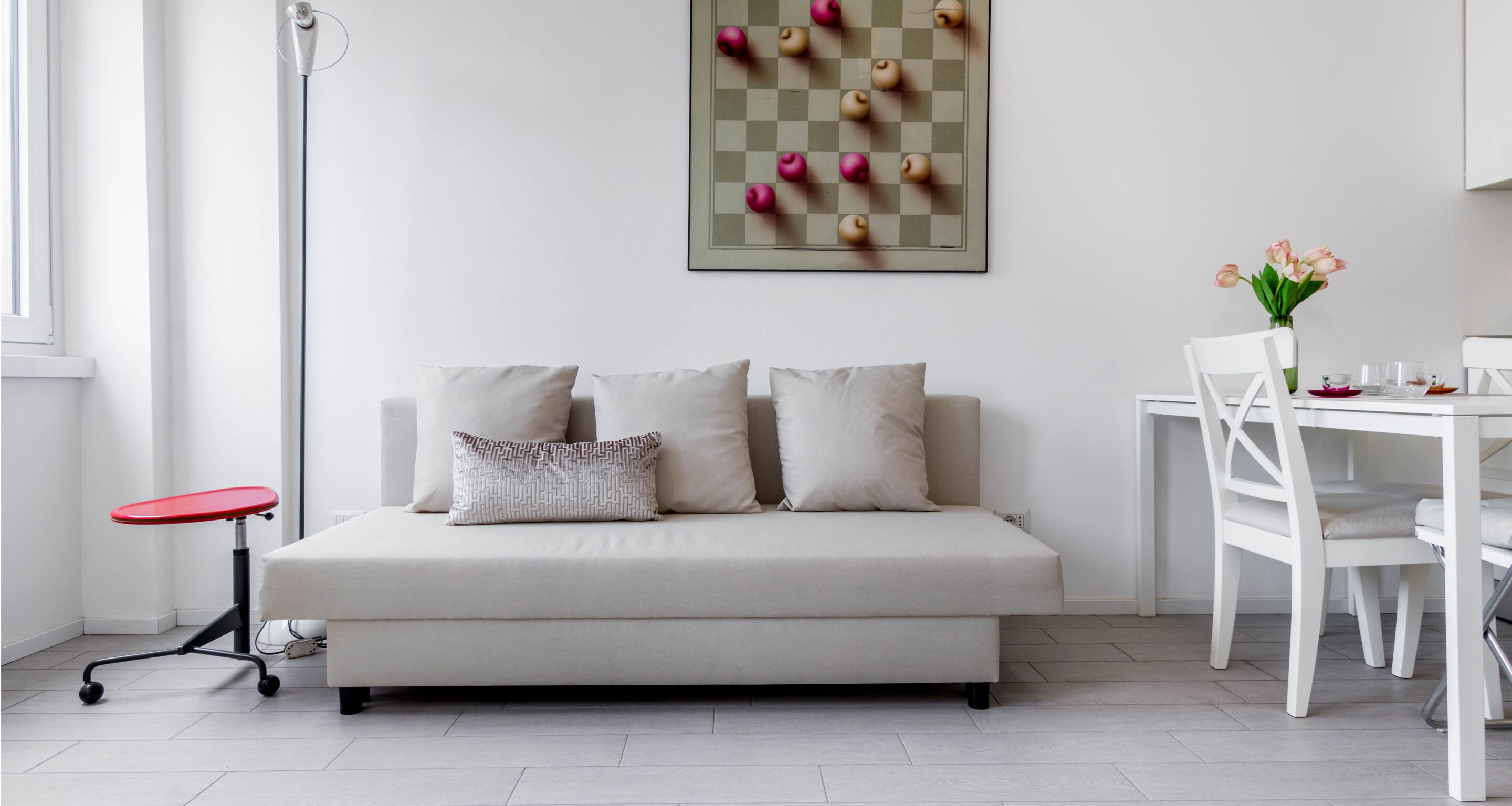 Un salon moderne avec un canapé, une petite table rouge, une table à manger blanche et une œuvre d’art accrochée au mur.