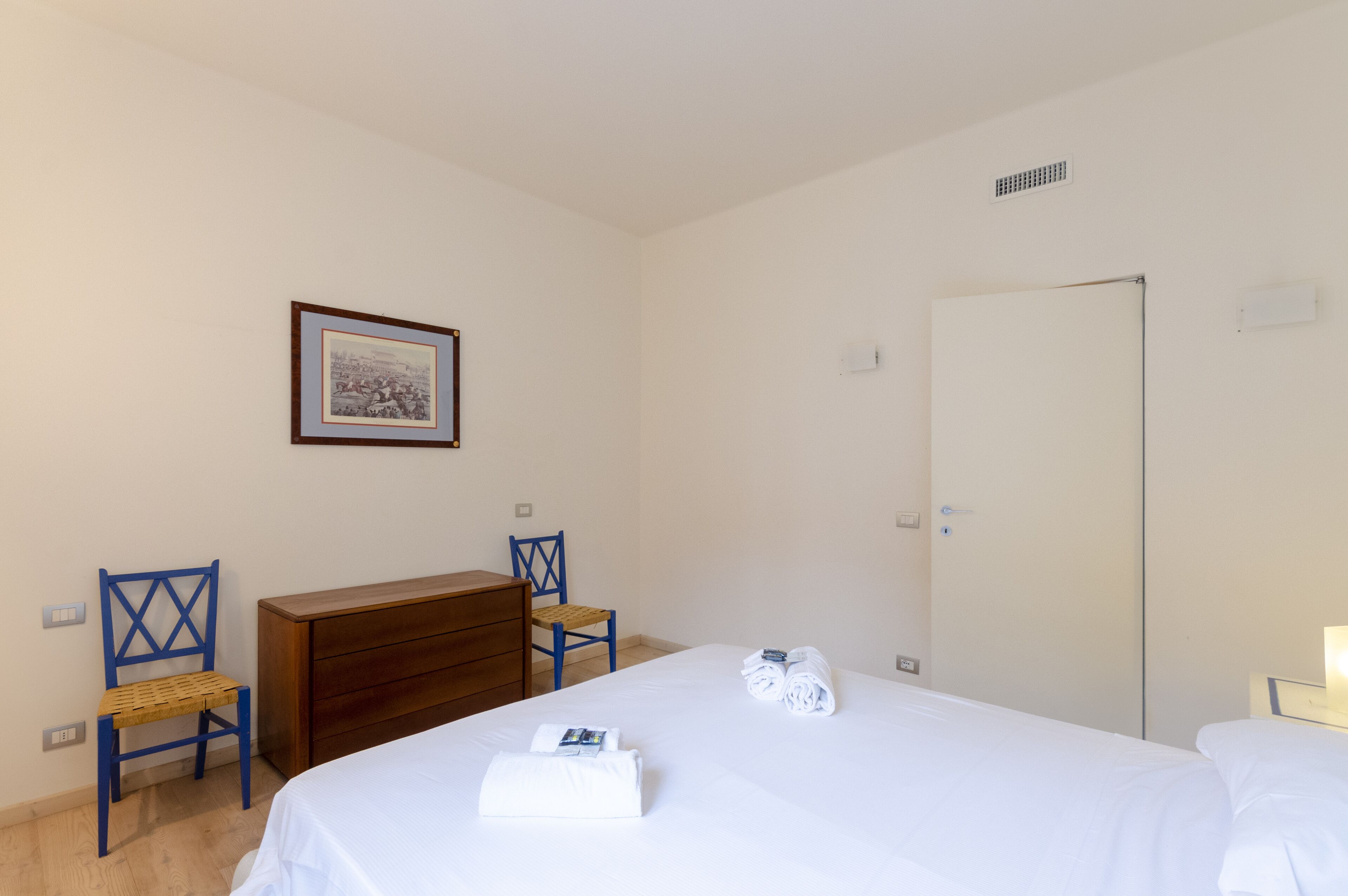 A hotel room with a bed, a wooden dresser, two blue chairs, a framed picture on the wall, and a white door.