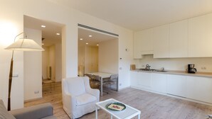 A modern living room with a sofa, a small table with a decorative plate, a dining area with chairs, and a kitchen with white cabinets and a coffee maker. - JOIVY Bright 3-bedroom apartment in Santa Margherita (Santa Margherita Ligure)