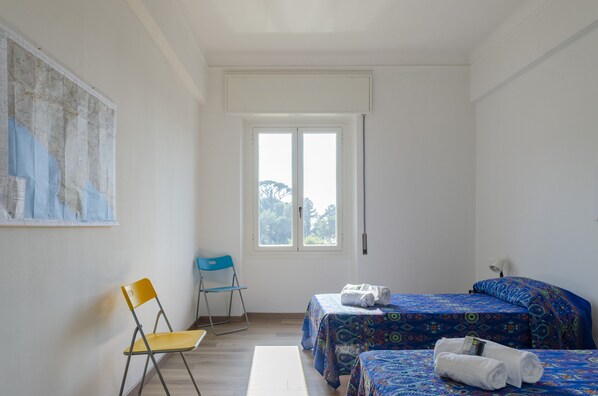 A room with two beds, a chair, a window, and a wall-mounted lamp.