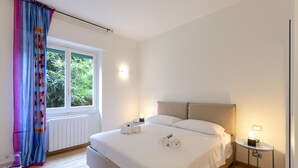 A neatly made bed with white linens, a bedside table with a lamp, and a window with curtains. - JOIVY 3-Bedroom Family Apartment in Santa Margherita near Beach (Santa Margherita Ligure)