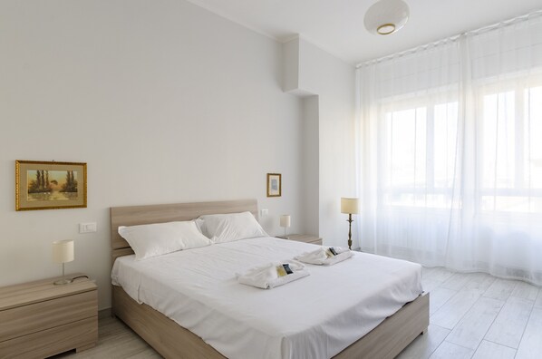 A hotel room with a large bed, two bedside tables with lamps, a ceiling light, and a window with sheer curtains.
