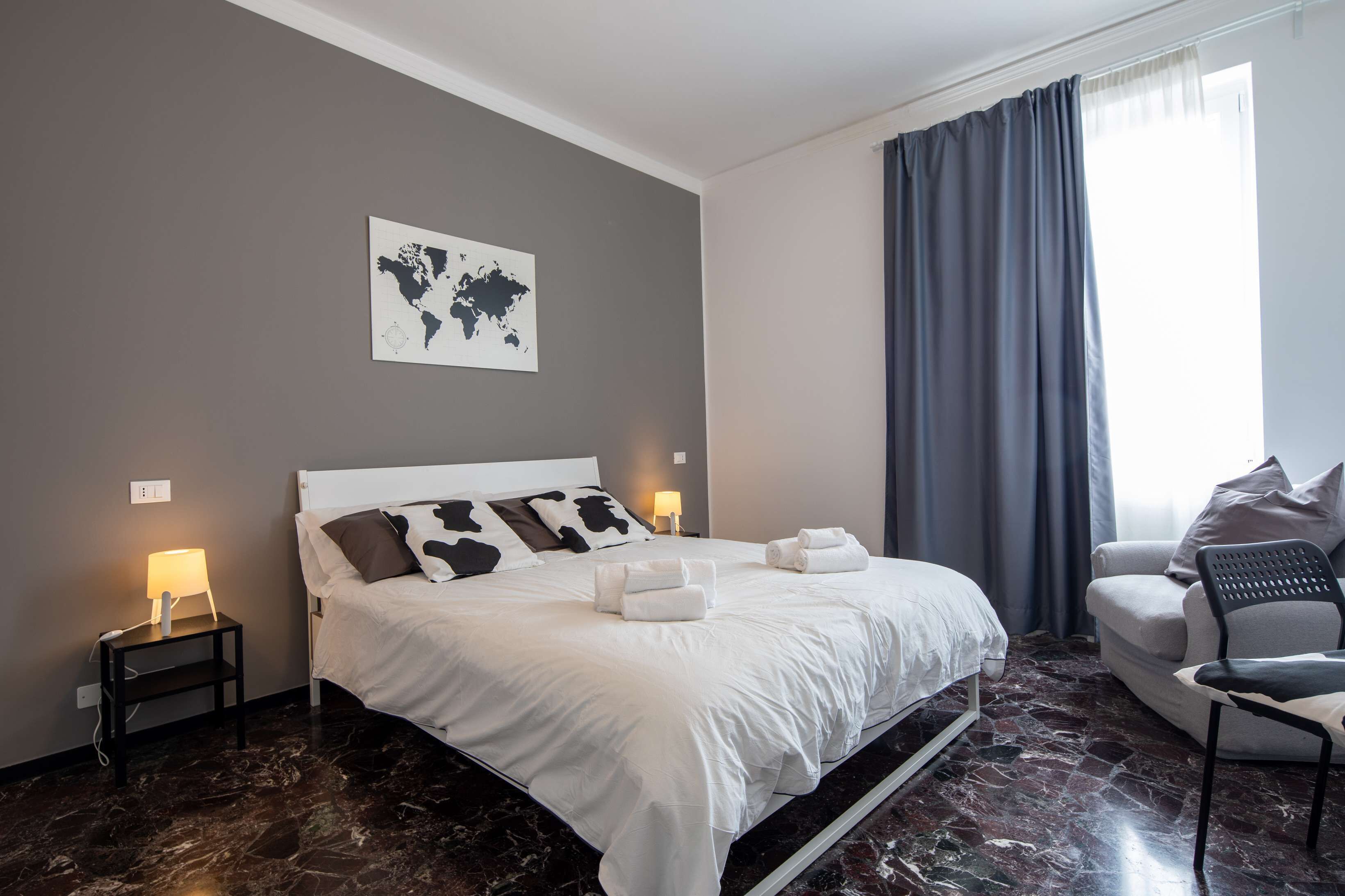 A neatly made bed with white linens and two pillows, a world map on the wall, a nightstand with a lamp, and a chair with a cushion.