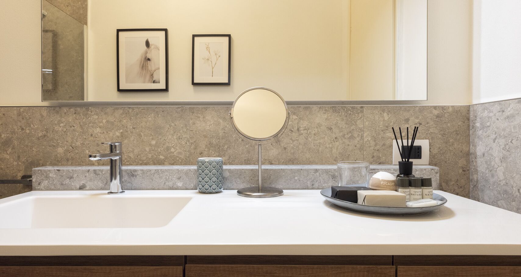 A bathroom with a large sink, a mirror, framed artwork on the wall, and toiletries on the counter.