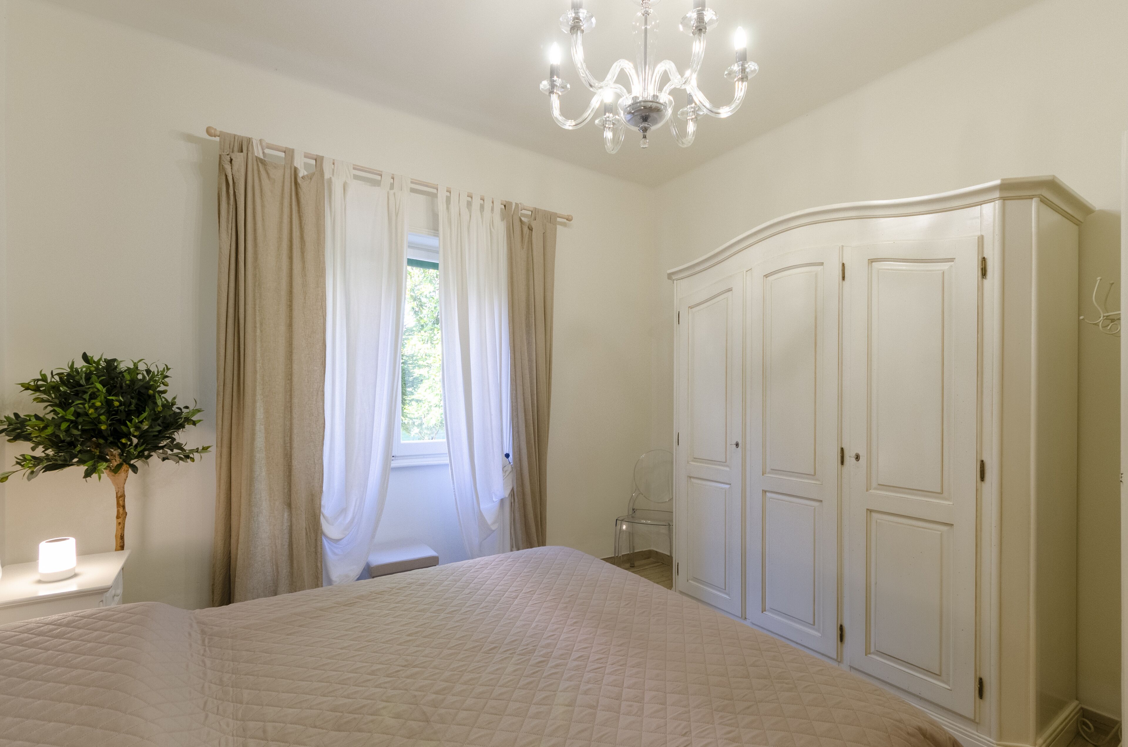 A bedroom with a bed, a wardrobe, a chandelier, and a window with curtains.